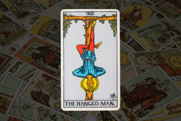 Montagem com a carta O Pendurado do Tarot na posição normal. Ao fundo, há várias cartas de Tarot.