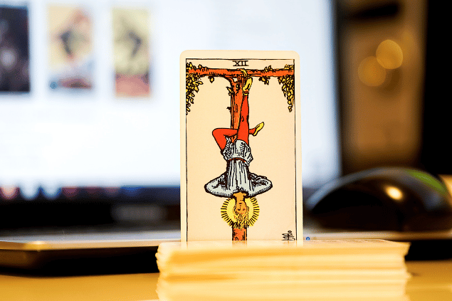 Na imagem, há a carta O Pendurado do Tarot na posição normal e em pé. Ao fundo, há um notebook aberto.
