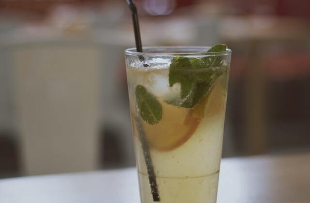 Copo transparente com líquido amarelo, folhas de hortelã e rodelas de limão com canudo preto.