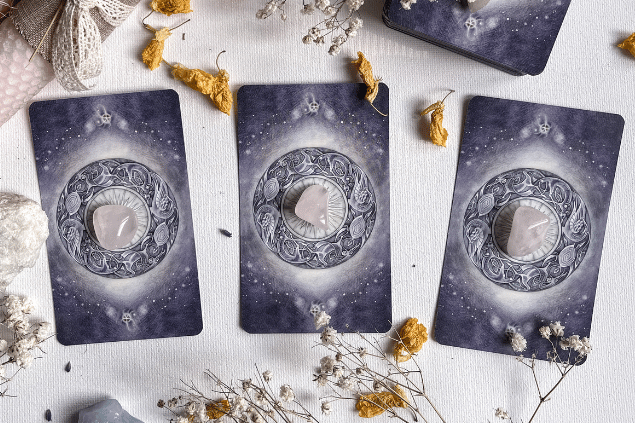 Três cartas de Tarot estão viradas para baixo em cima de uma mesa. Há um cristal rosa sobre cada uma dessas cartas. Ao lado, há flores e pedras.