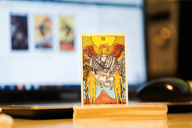 Na imagem, há a carta Os Enamorados do Tarot na posição normal e em pé. Ao fundo, há um notebook aberto.