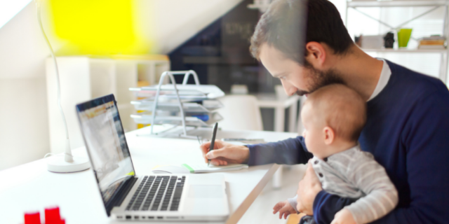 Pai segura bebê enquanto escreve em bloco de notas. Ele está sentado à mesa, onde há um computador.