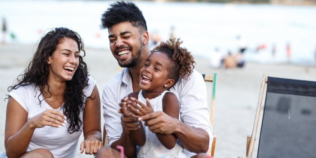 Homem e mulher adultos sentados em cadeiras de praia. Sobre as pernas do homem, há uma menina. Os três sorriem e estão em uma praia.