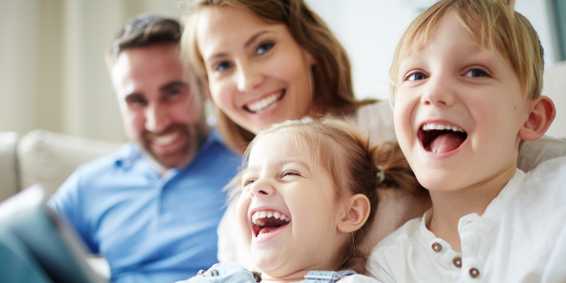 Família composta por duas crianças e dois adultos. Todos sorriem e estão abraçados.