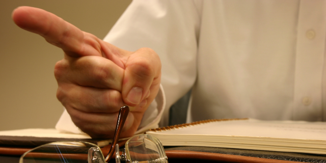 Homem com o dedo indicador erguido. Sua mão está apoiada sobre um caderno aberto; à frente, há óculos.