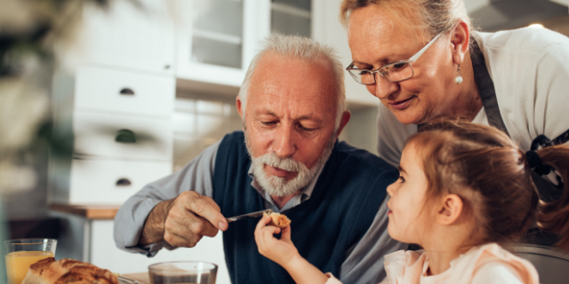 Avô e neta sentados à mesa; ele a ajuda a comer. Atrás de ambos, há a avó.