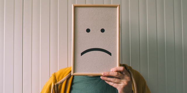 Homem segura quadro com representação de rosto triste sobre a sua própria face. Ao fundo, há cenário branco com elevações.