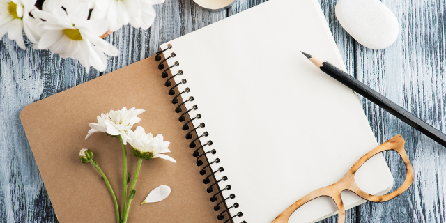 Diário aberto sobre mesa; na capa há flores brancas e, sobre a primeira página, uma armação de óculos e um lápis.