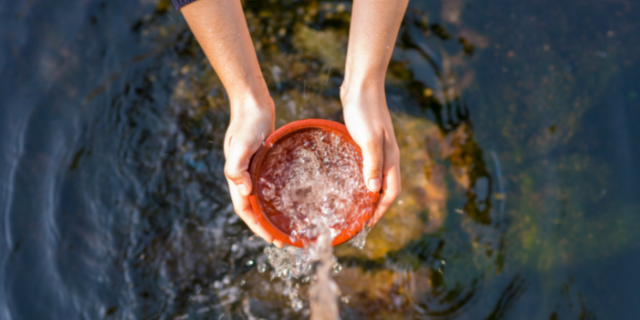 Água cai em recipiente marrom segurado por duas mãos. Ao fundo, há um lago.