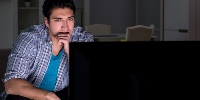 Homem sentado em frente à uma televisão. Ele está com um semblante concentrado e tem a mão sobre a boca, além de apoiar os cotovelos nos joelhos.