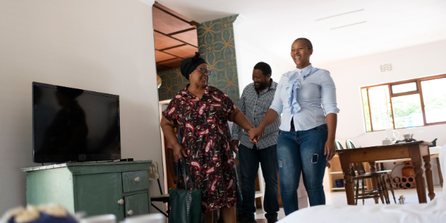 Em uma sala de estar, duas mulheres de mãos dadas caminham para a frente. Ao fundo, há um homem rindo e olhando para baixo.