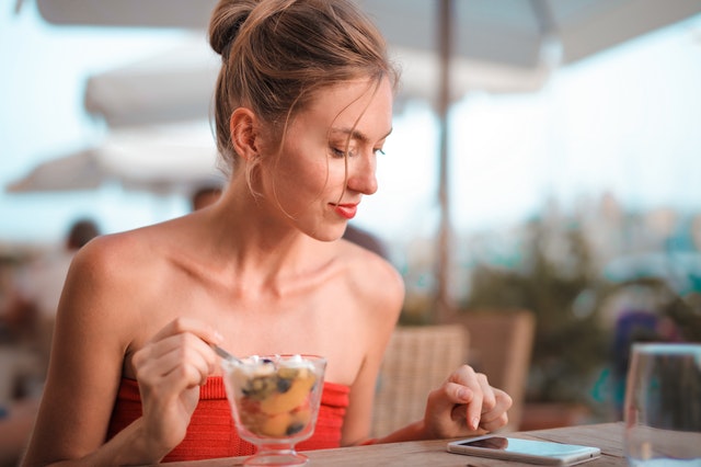 Mulher comendo uma salada de frutas em um restaurante enquanto usa o celular.