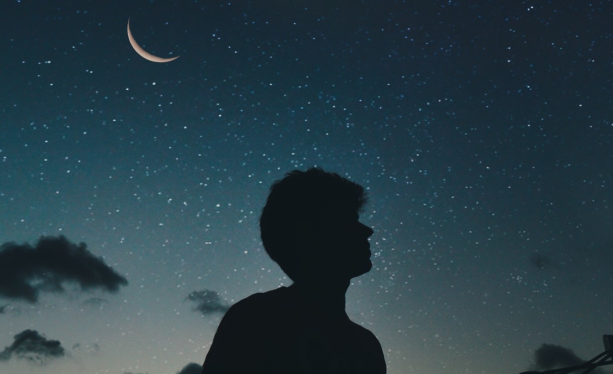 Silhueta de um homem com o céu estrelado de fundo e a lua posicionada à esquerda 
