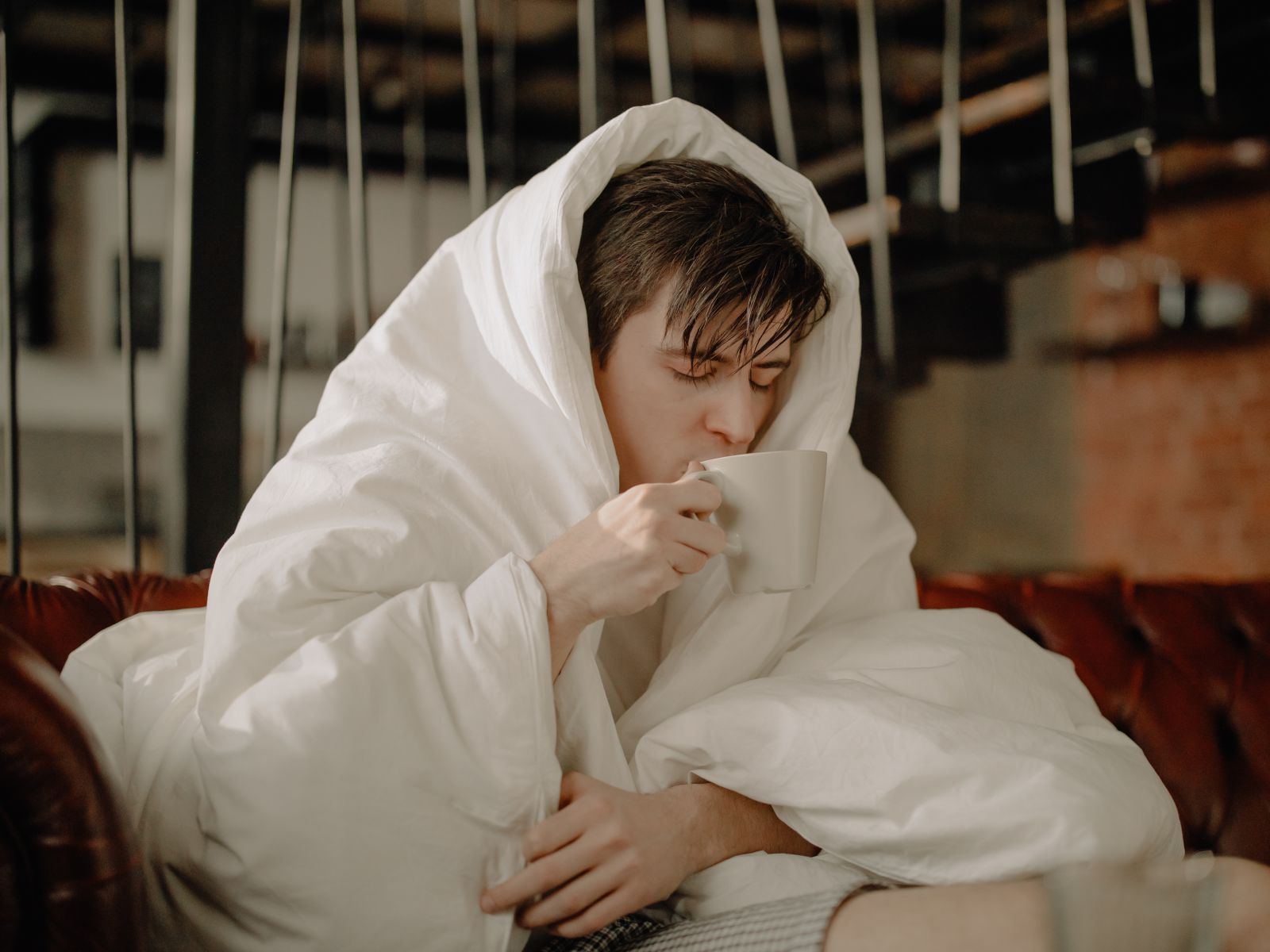 Homem sentado em um sofá enrolado em um cobertor e segurando uma xícara