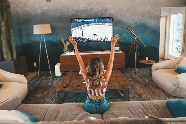 Mulher branca sentada num sofá com os braços levantados enquanto assiste um jogo de hóquei,