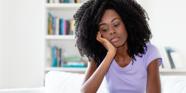 Mulher cabisbaixa apoia o rosto sobre a mão. Ela está sentada e, ao fundo, há uma estante com livros. 