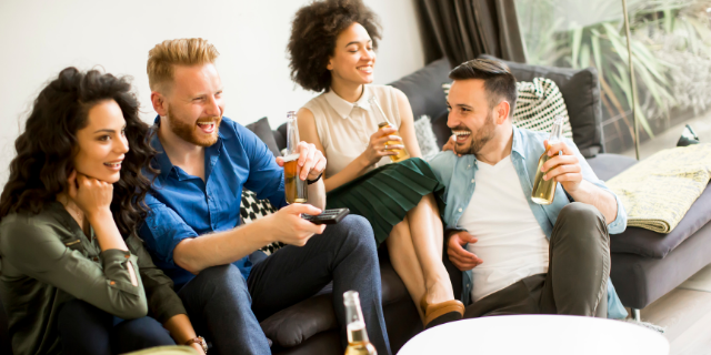 Grupo de amigos em sala de estar. Eles sorriem e bebem cervejas. Um deles segura um controle remoto.