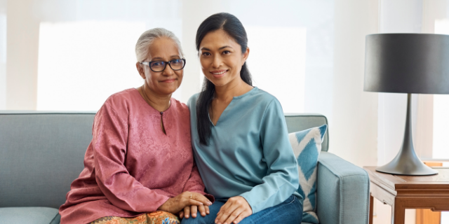 Duas mulheres, uma mais jovem e outra mais velha, sentadas lado a lado em um sofá. O cenário é uma sala de estar.