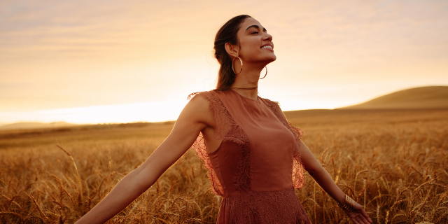 Mulher em campo sorri e abre os braços; seus olhos estão fechados.