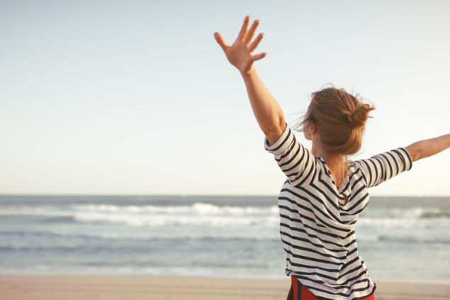 Mulher de costas, com os braços abertos em direção ao mar.