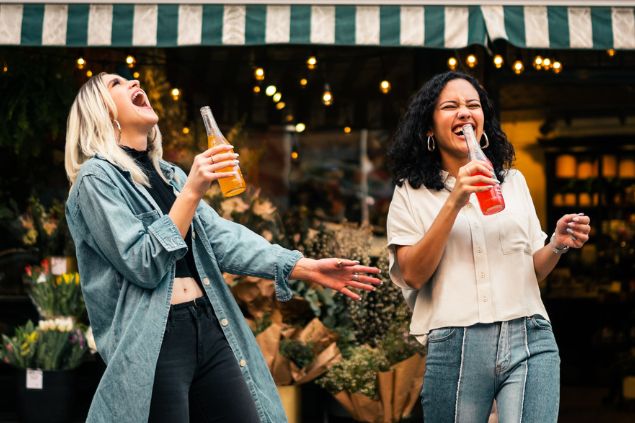 Duas amigas rindo alegremente e bebendo suco na rua.