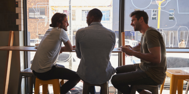 Três homens sentados à mesa sorriem e conversam. 