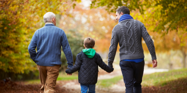 Um menino anda entre dois homens. Eles estão de mãos dadas e caminham por um cenário externo.