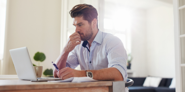 Homem sentado à mesa, onde há um computador e papéis. 