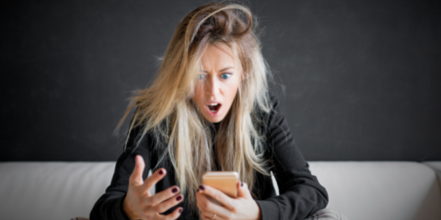 Mulher com semblante surpreso encara um celular que segura em uma das mãos. Ela está sentada em uma cama.