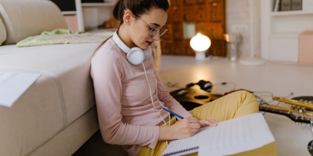 Mulher sentada no chão apoiada em sofá branco. Ela está com fones pendurados no pescoço e escreve em um caderno apoiado sobre sua perna. Ao seu lado, há um violão sobre o chão.