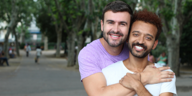 Casal em ambiente externo. Um homem abraça outro que está em sua frente.