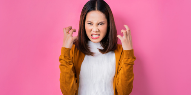 Mulher com as mãos erguidas próximas ao rosto. Seu semblante é de raiva.
