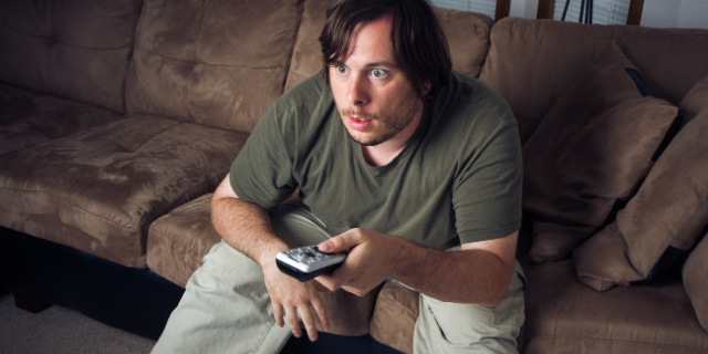 Homem com um semblante surpreso está sentado em um sofá. Ele segura um controle remoto e apoia os cotovelos nos joelhos.
