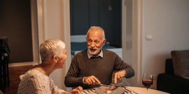 bernardbodo / Canva Casal janta juntos à mesa e sorri.
