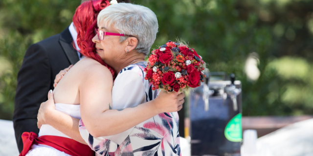 Duas mulheres, sogra e nora, abraçam-se em cerimônia de casamento. Ao fundo, há um homem vestido socialmente. O cenário é externo e arborizado.
