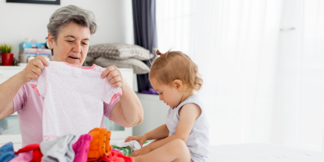 Avó segura roupa infantil em suas mãos. Em sua frente, há uma cama com mais roupas e uma bebê.