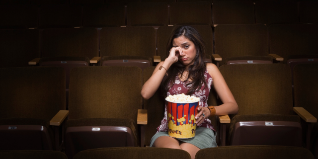 Mulher em cinema chora. Ela segura um balde de pipoca.