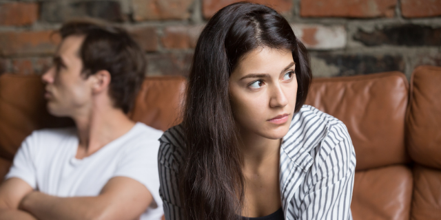 Casal sentado em um sofá. O homem, ao fundo, está com o rosto virado ao lado oposto da parceira, senta à frente. 
