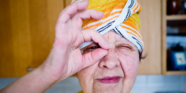 Idosa faz gesto circular com o dedo indicador e polegar ao redor do olho direito. Ela está de turbante.