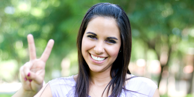 Mulher sorri e simboliza paz e amor com uma de suas mãos.